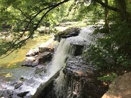Water falling off rocks into a river. Image taken from a biking trail. There are green trees all around. 