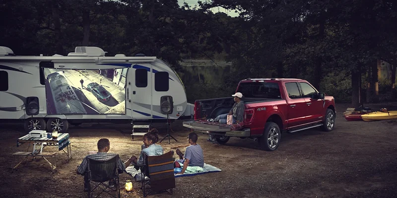 Family who took out the new 2023 Ford F-150 out camping with their travel trailer; sitting around a camp fire and watching a movie projected onto the side of the travel trailer.