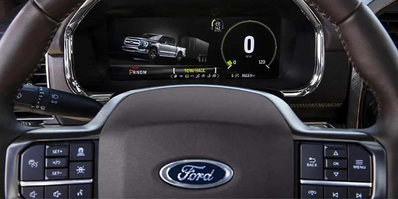 close up shot of a 2023 Ford F-150's steering wheel looking through it at the speedometer area that shows a Ford F-150 towing. You can see the speedometer section, drive section, and tow/haul options section.