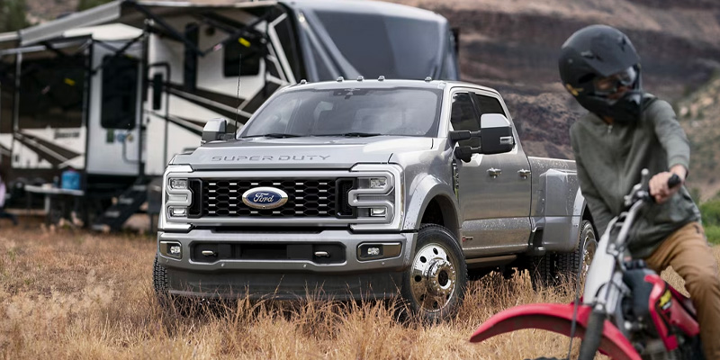 A silver 2025 Ford F-250 Super Duty parked on grass with a camper in the background and a motocross rider nearby.